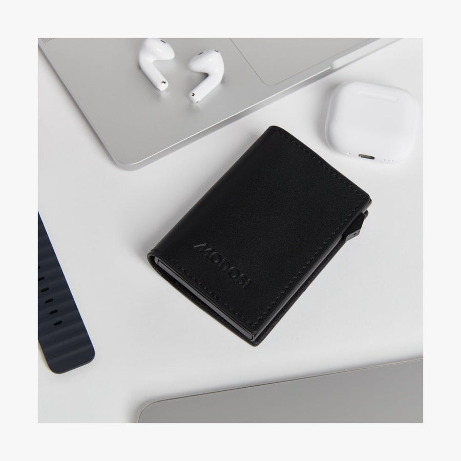 Carbon Black / Black | Slim Wallet Pro in Carbon Black / Black placed on a desk alongside wireless earbuds, a laptop, and a smartwatch band, highlighting its sleek and modern design in a lifestyle setting.