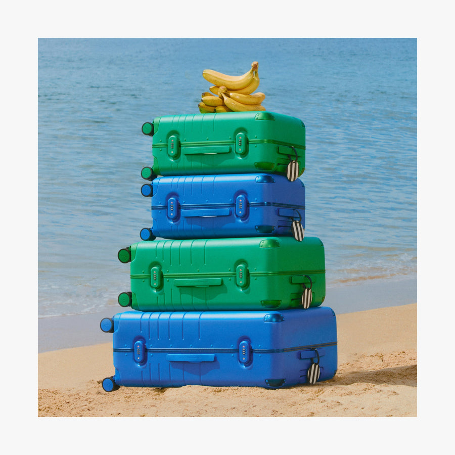 Jungle Green | Stack of Jungle Green and Coastal Blue Hybrid suitcases in different sizes with a banana on top, set on a sandy beach