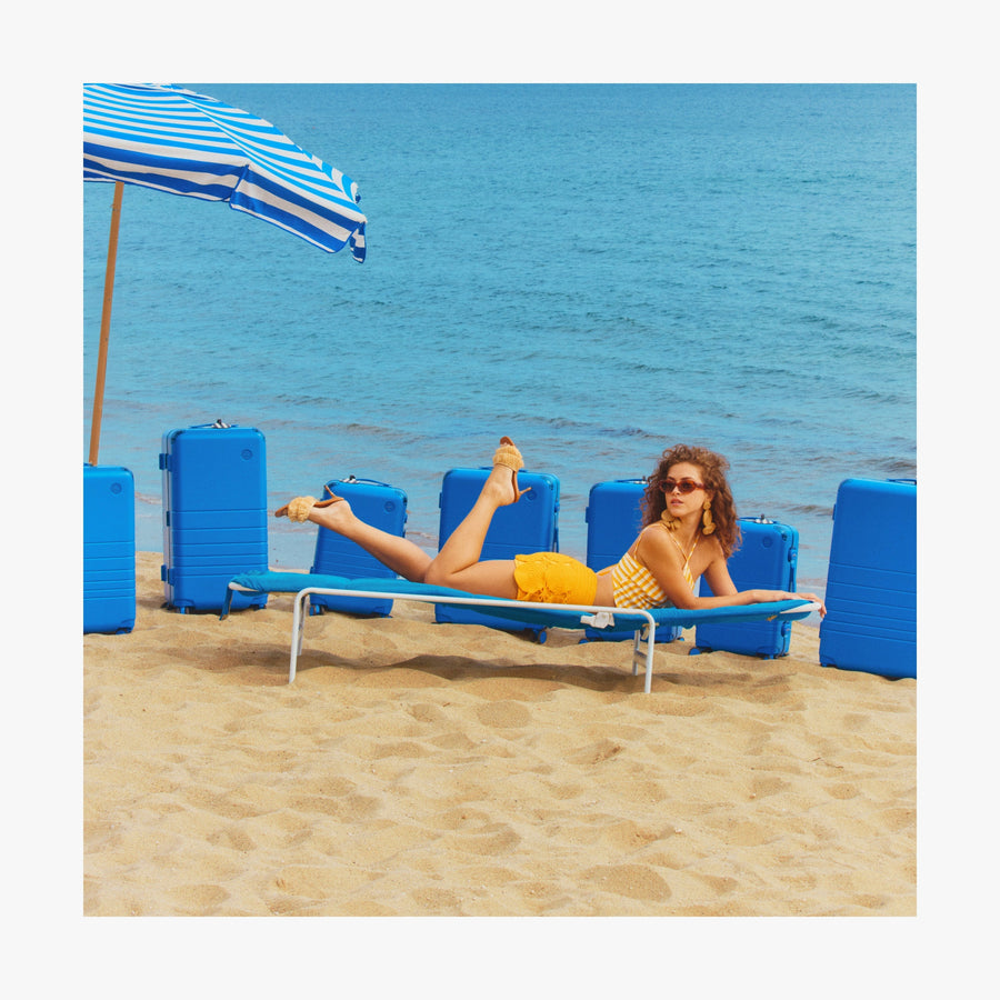Coastal Blue | Woman lounging on a beach chair under a striped umbrella, with Coastal Blue Hybrid suitcases arranged behind her on the sand by the ocean