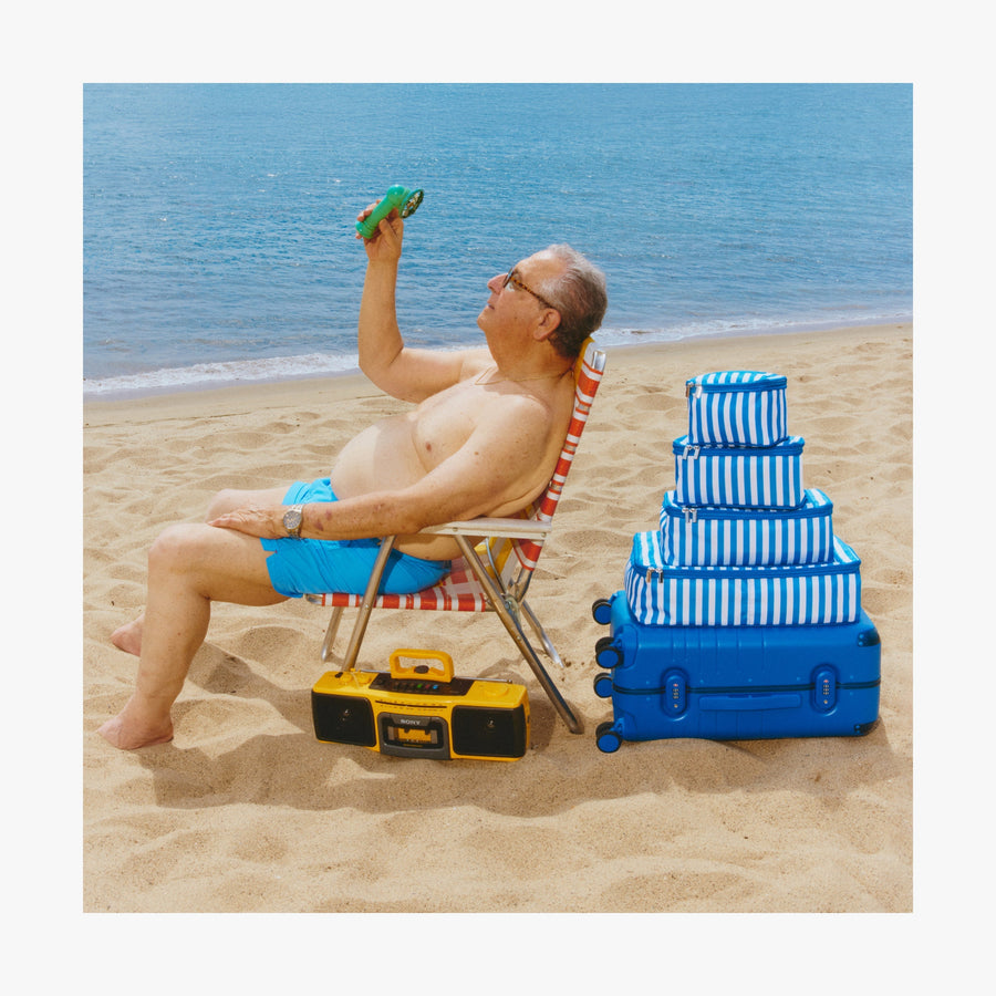 Set of Six / Poplin Stripe Coastal Blue | Man relaxing on a chair, cooling off with a portable fan beside a stack of Coastal Blue Hybrid suitcases and Poplin Stripe Coastal Blue Packing Cubes in various sizes