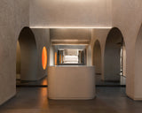 The reception area of the Monos Georgetown store, framed by textured stone arches and softly glowing circular light features. A curved minimalist counter stands at the center, reflecting the warm tones of the surrounding mirrors and brushed steel surfaces.
