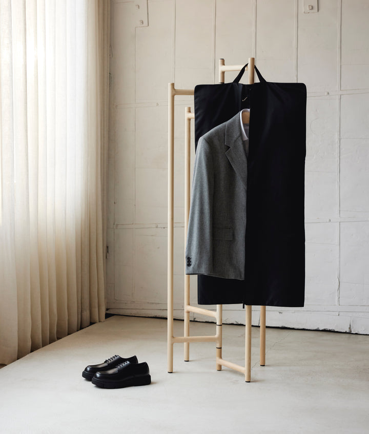 The Monos Garment Sleeve hanging on a minimalist wooden rack beside a tailored grey blazer and black loafers. A refined travel essential designed to keep formalwear crisp and organized, captured in a serene, light-filled room.