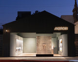Exterior view of the Monos Abbot Kinney store in Los Angeles at dusk. The modern black façade features a softly glowing Monos sign above the entrance. Warm interior lighting highlights a curved glass block wall and minimalist display shelves on each side.