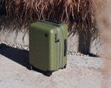Monos Carry-On in Olive outdoors on sunlit concrete with dry hanging plants behind.